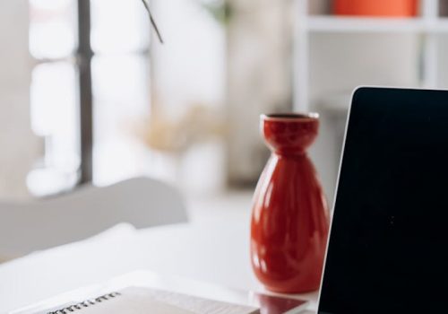 A minimalist home office setup featuring a laptop, notebook, and a red vase on a white desk.